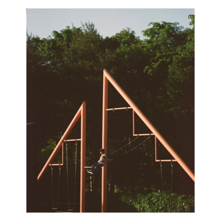 Child swings on a pink metal swing set, surrounded by lush green trees under a bright sky. Outdoor playground in a park setting.