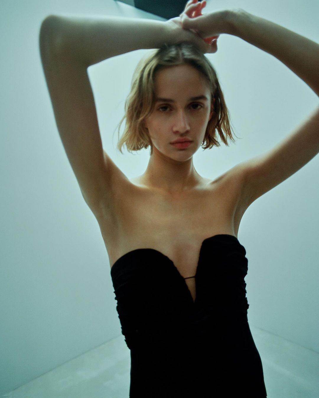 Young woman in a strapless black dress poses with hands up, shot from a low angle against a pale cyan studio backdrop.