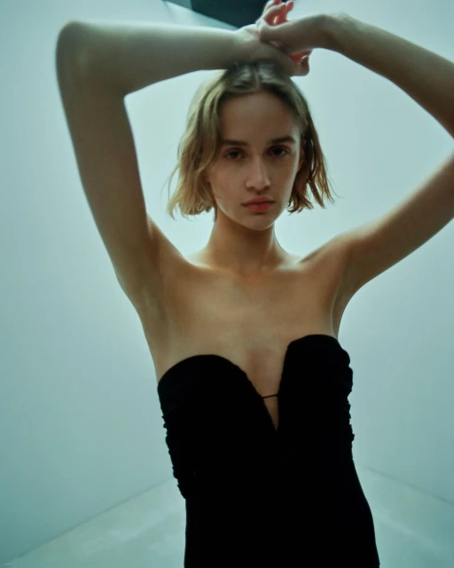 Young woman in a strapless black dress poses with hands up, shot from a low angle against a pale cyan studio backdrop.