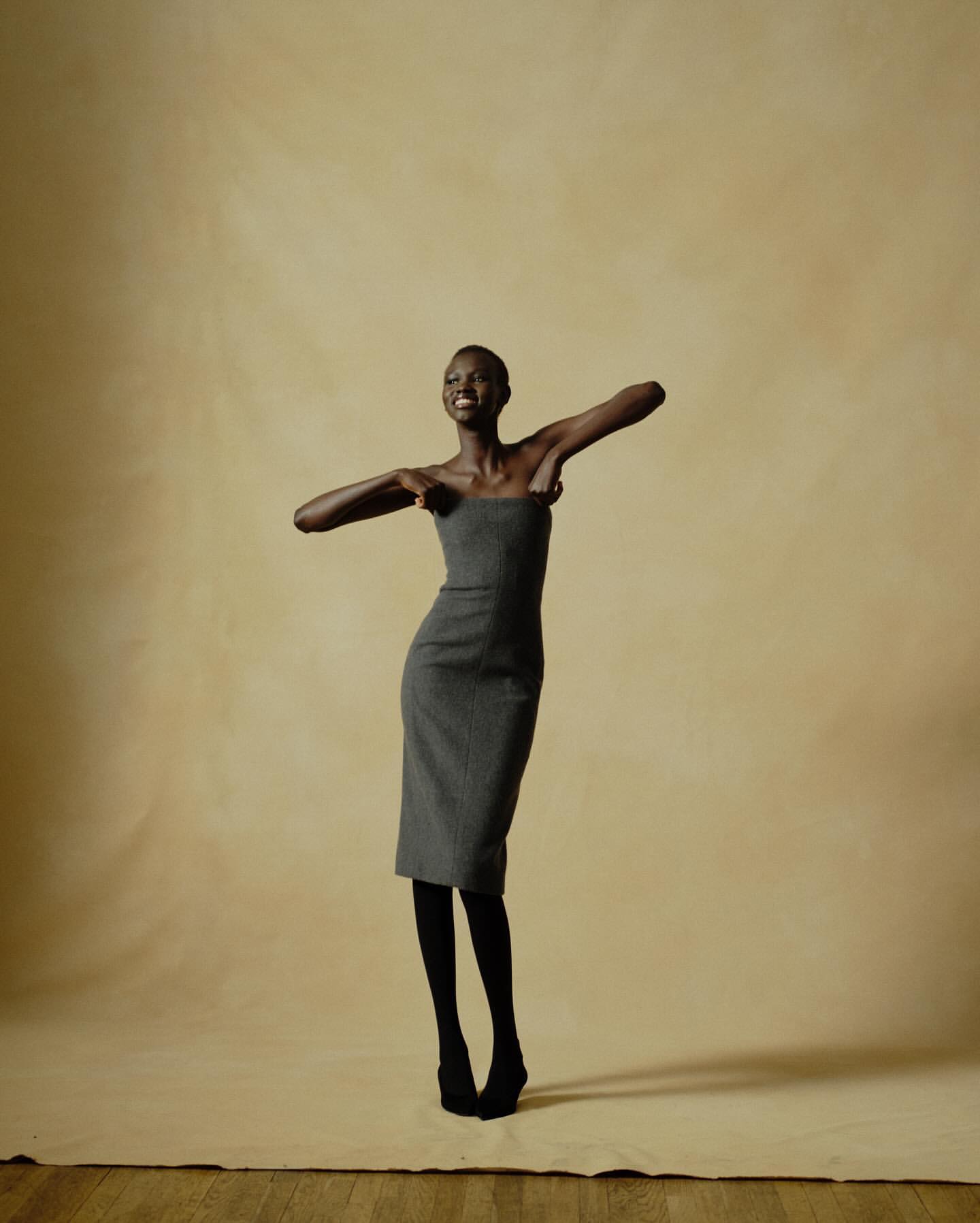 A tall, slender Black woman in a gray strapless dress smiles in a studio setting, complemented by black tights and shoes against a muted backdrop.