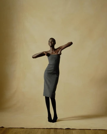 A tall, slender Black woman in a gray strapless dress smiles in a studio setting, complemented by black tights and shoes against a muted backdrop.