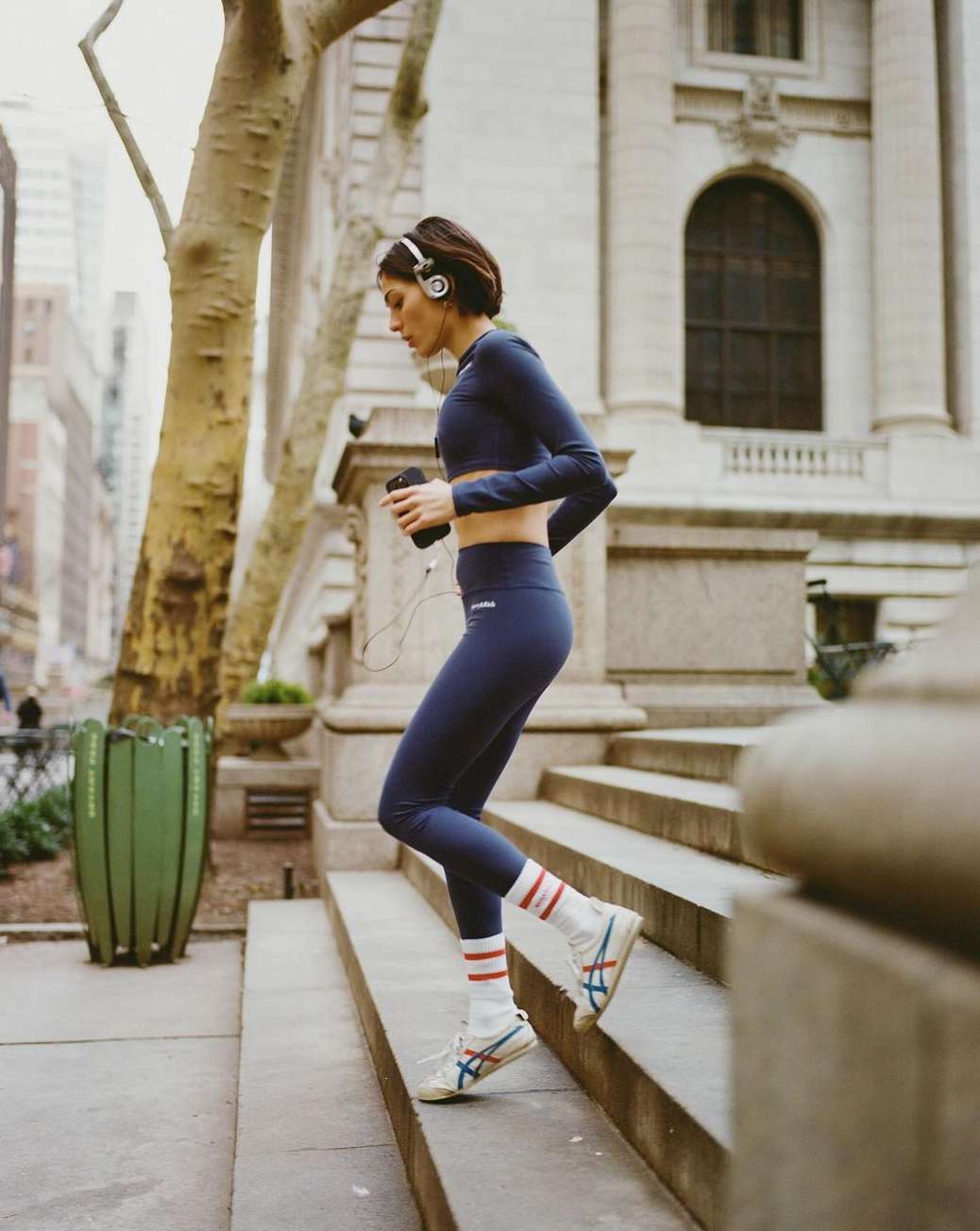 Athletic woman in sportswear climbs outdoor steps in city; wearing headphones, holding a phone, with trees and buildings in the background.