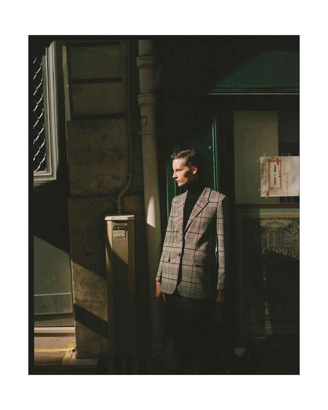 Stylish male model in a plaid blazer stands in an urban alleyway, bathed in sunlight, for a fashion editorial shoot.