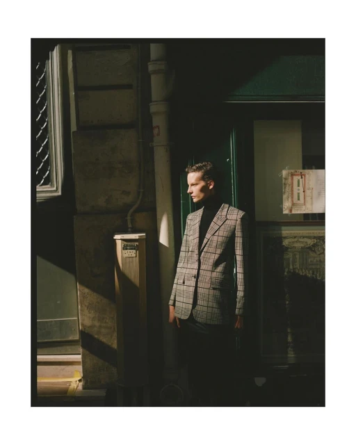 Stylish male model in a plaid blazer stands in an urban alleyway, bathed in sunlight, for a fashion editorial shoot.
