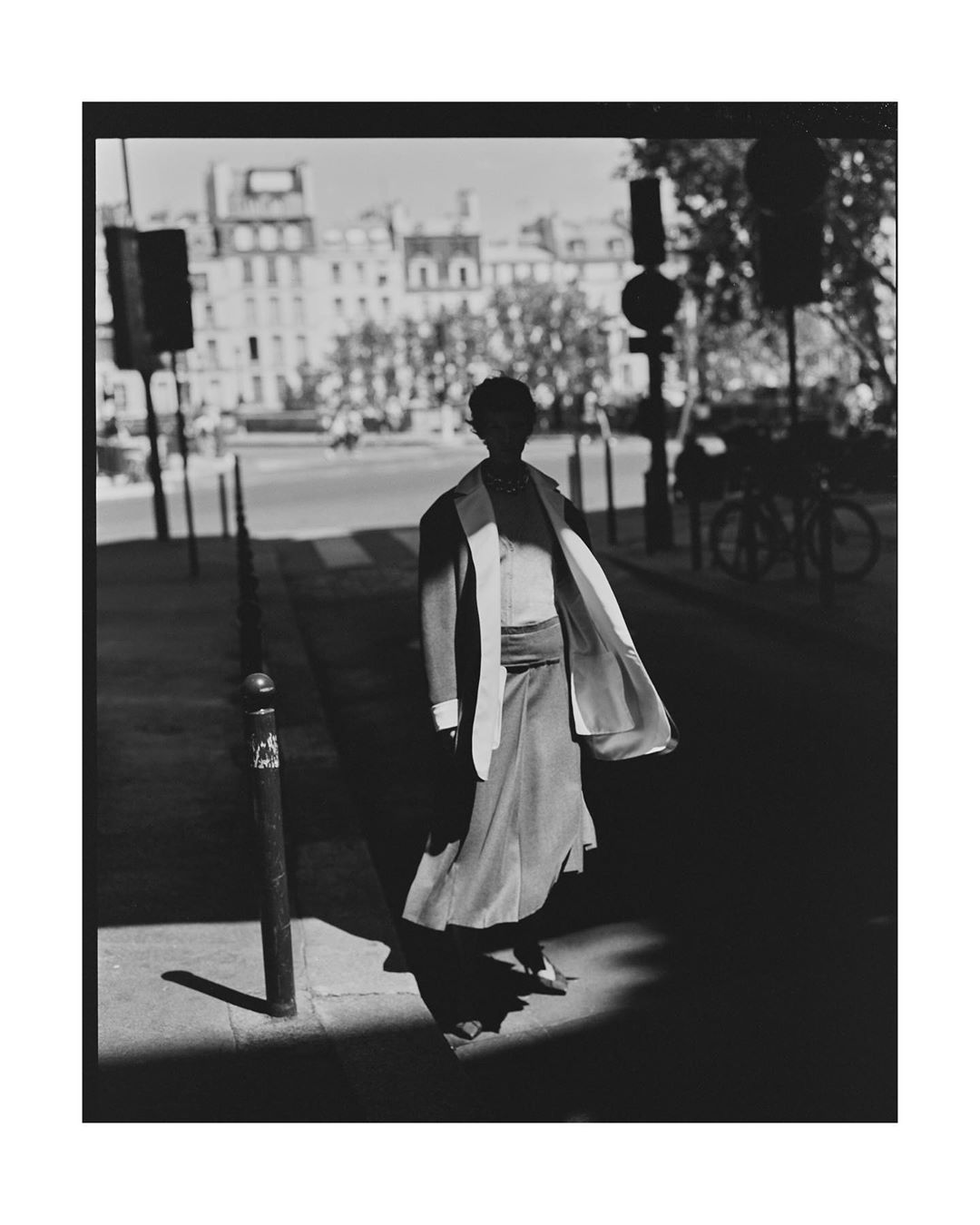 Black and white street style photo of a woman in a skirt and blazer walking on a city street with buildings and trees in the background.