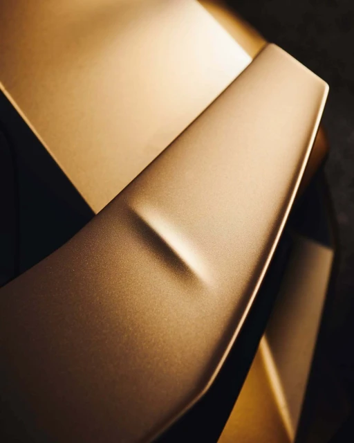 Close-up of a golden-colored metallic car spoiler with a matte finish, reflecting light in a studio setting.