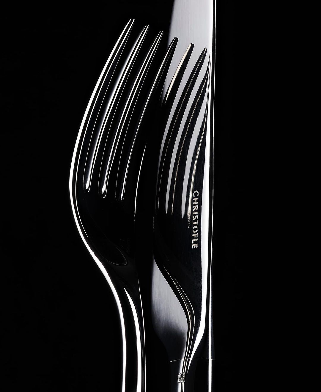 Close-up studio shot of a silver Christofle knife and fork. The lighting is high contrast and emphasizes the shiny metal of the utensils.
