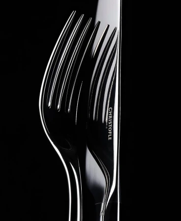 Close-up studio shot of a silver Christofle knife and fork. The lighting is high contrast and emphasizes the shiny metal of the utensils.