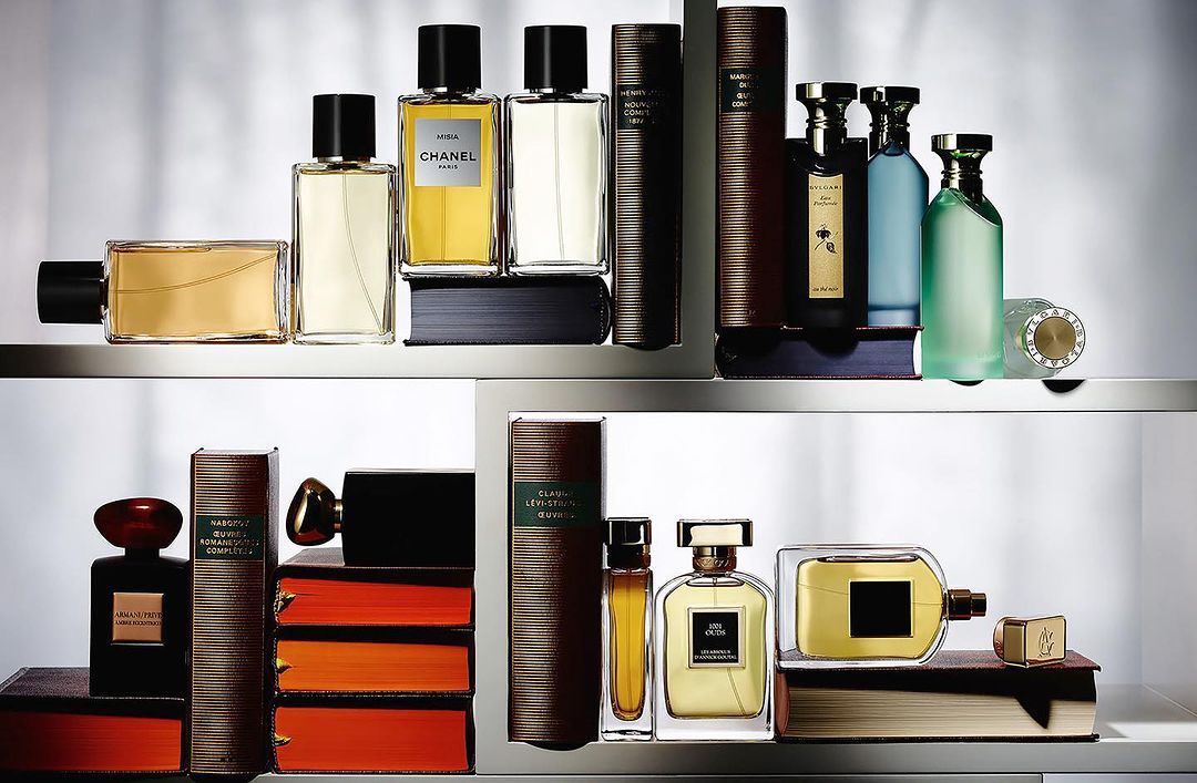 Still life of multiple perfume bottles of different shapes and colors displayed on shelves with books, under bright lighting.