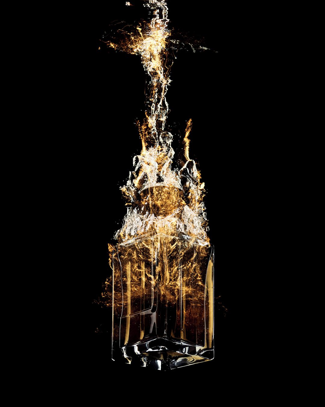 A clear glass bottle with amber liquid inside is engulfed in splashing water against a stark black background.