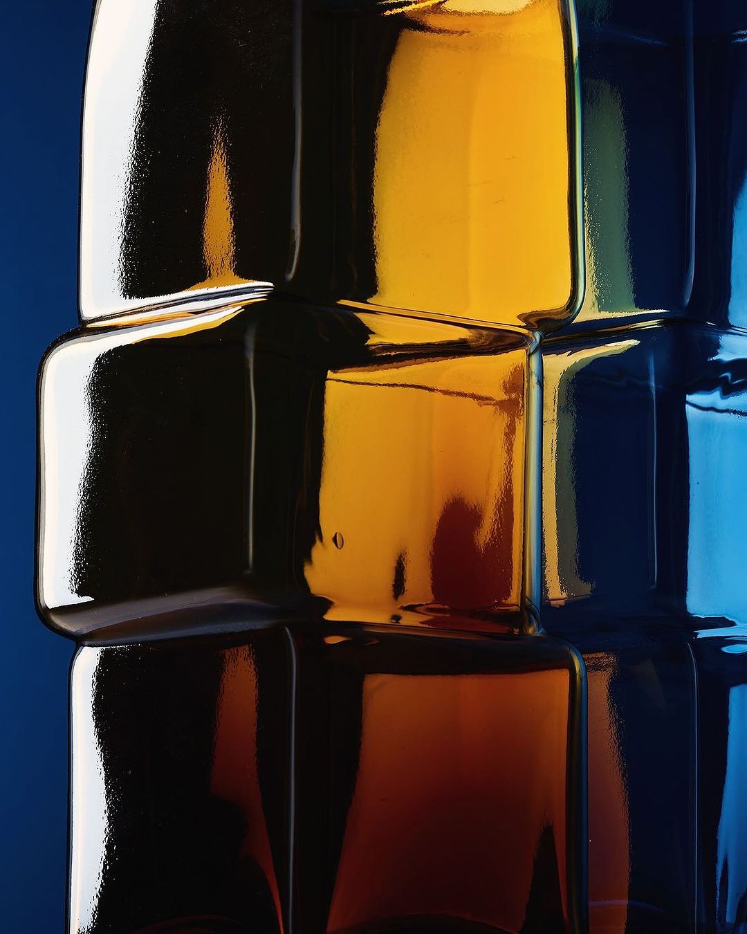 Abstract stacked square bottles in shades of blue, brown, and yellow arranged against a deep blue backdrop in vertical orientation.