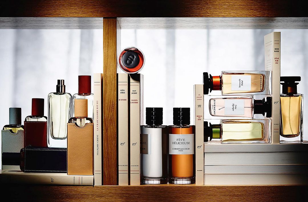 Still life shot of an arrangement of assorted designer perfume bottles and hardcover books on a wooden shelf against a bright background.