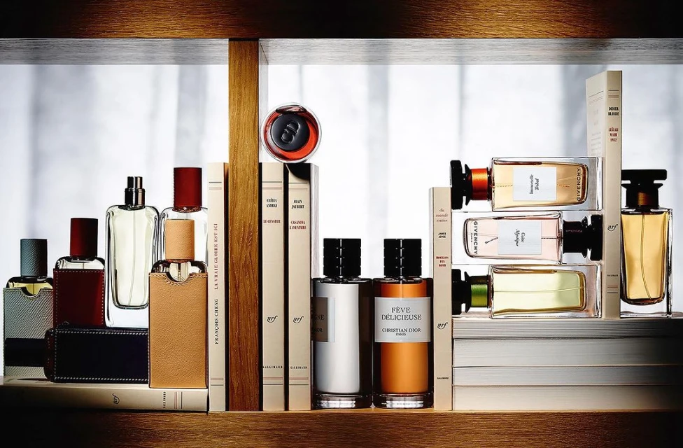 Still life shot of an arrangement of assorted designer perfume bottles and hardcover books on a wooden shelf against a bright background.