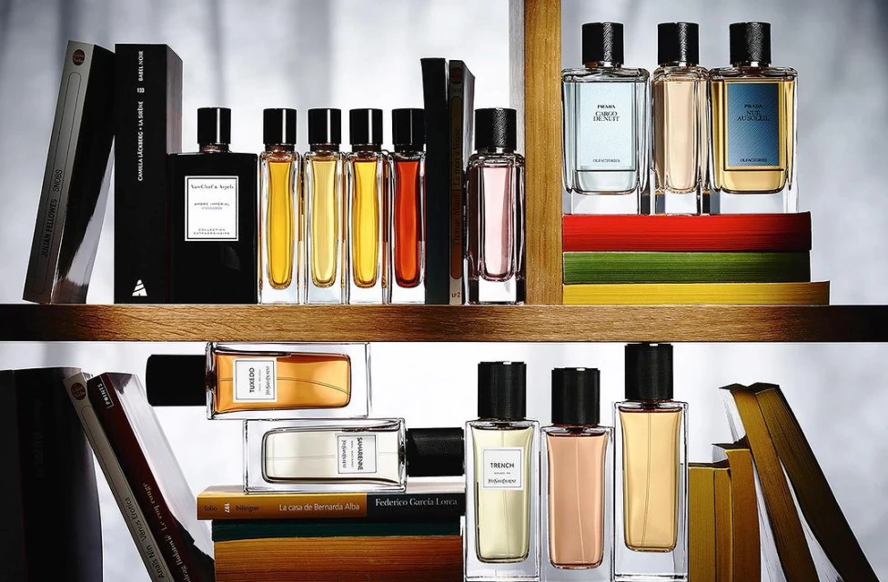 Various glass perfume bottles on wooden shelves with hardback books. The bottles are filled with liquids of varying colors from clear to amber.