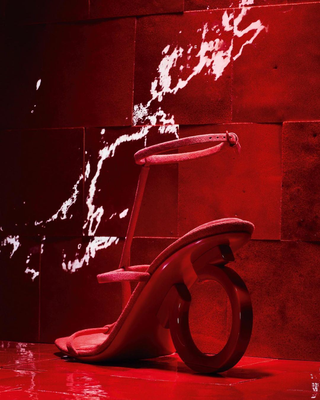Striking studio shot of a modern, red-toned sandal with a round heel, displayed against a textured red wall with a splash of white lighting.
