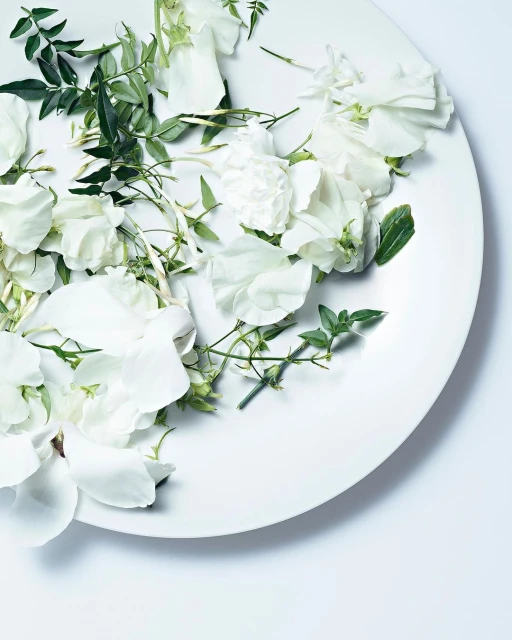 Overhead view of a white plate filled with white flowers and green leaves on a white background, creating a clean and floral aesthetic.