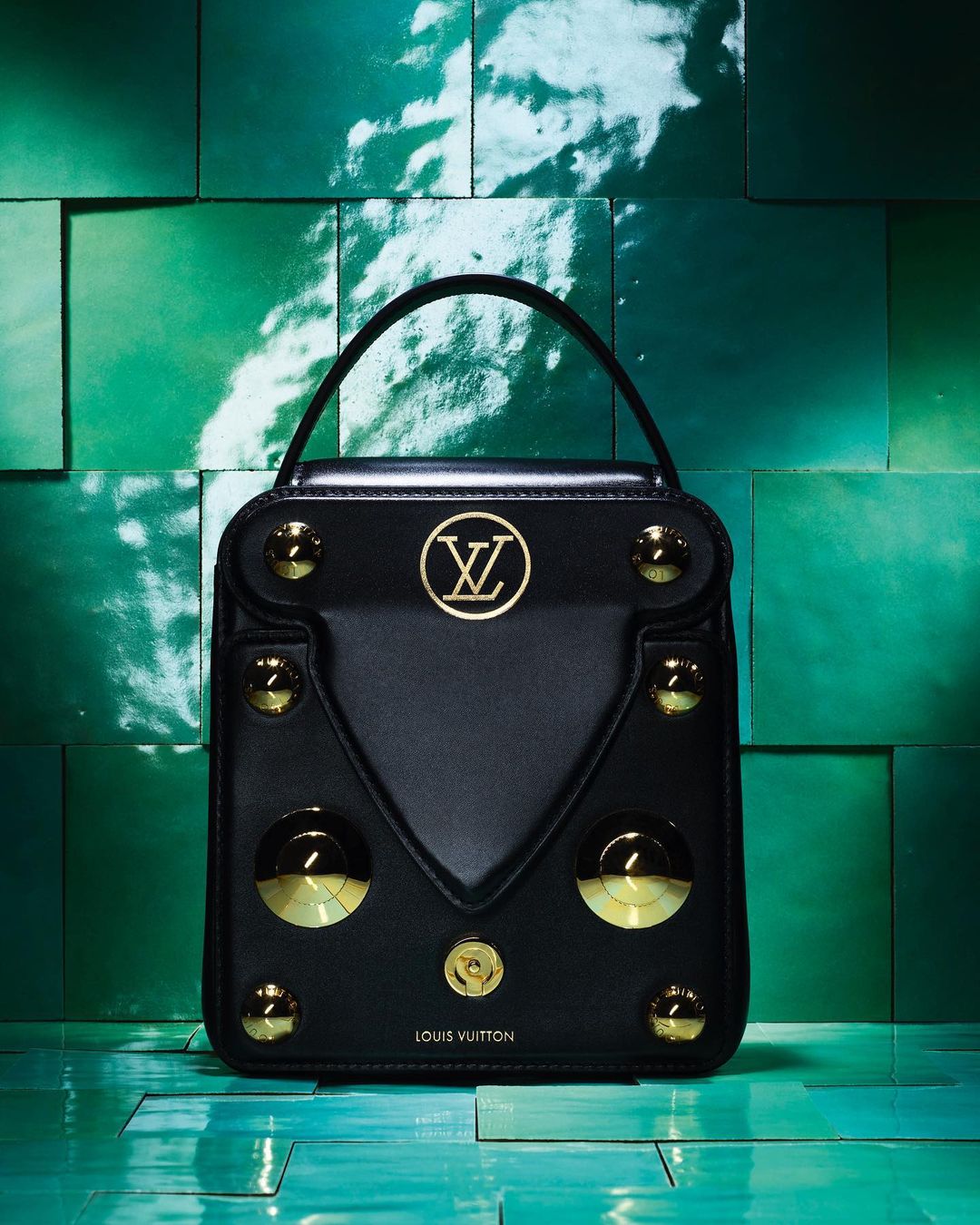 Black Louis Vuitton top-handle bag featuring gold studs and logo on a glossy teal tile background, capturing luxury and brand recognition.