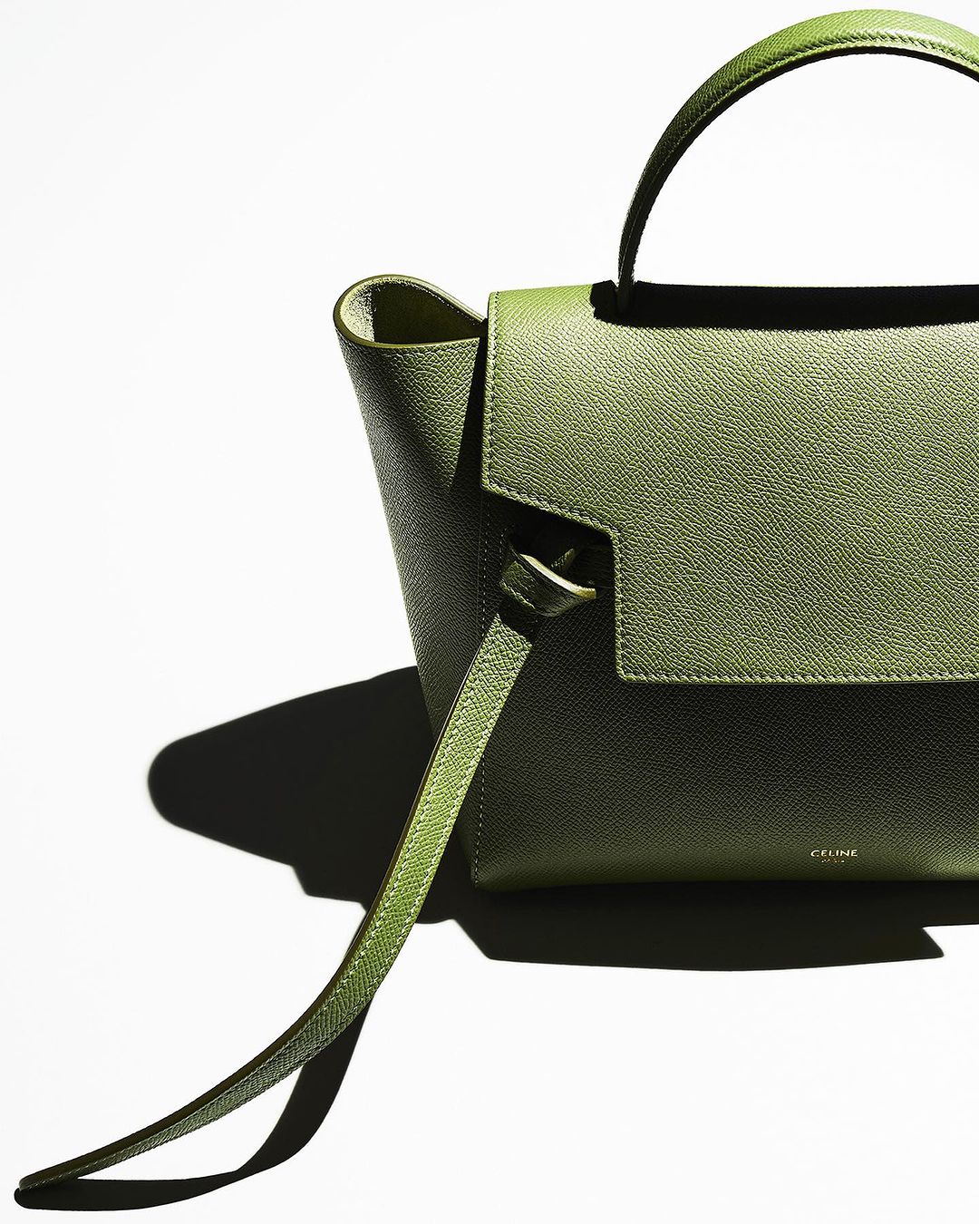 Close-up of an olive-green leather handbag with a top handle and shoulder strap against a white backdrop, emphasizing its texture and design.