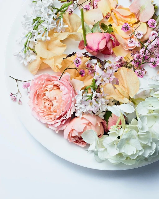 Overhead shot of white plate covered with pastel roses, lilacs, hydrangea, and petals in a soft, elegant floral arrangement.