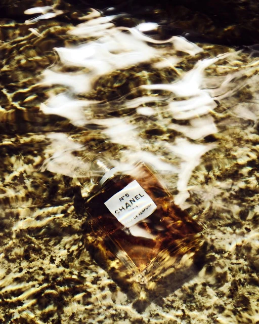 Chanel No. 5 perfume bottle submerged in shallow, clear water with rippling surface and a brown and white sandy bottom visible.