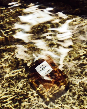 Chanel No. 5 perfume bottle submerged in shallow, clear water with rippling surface and a brown and white sandy bottom visible.