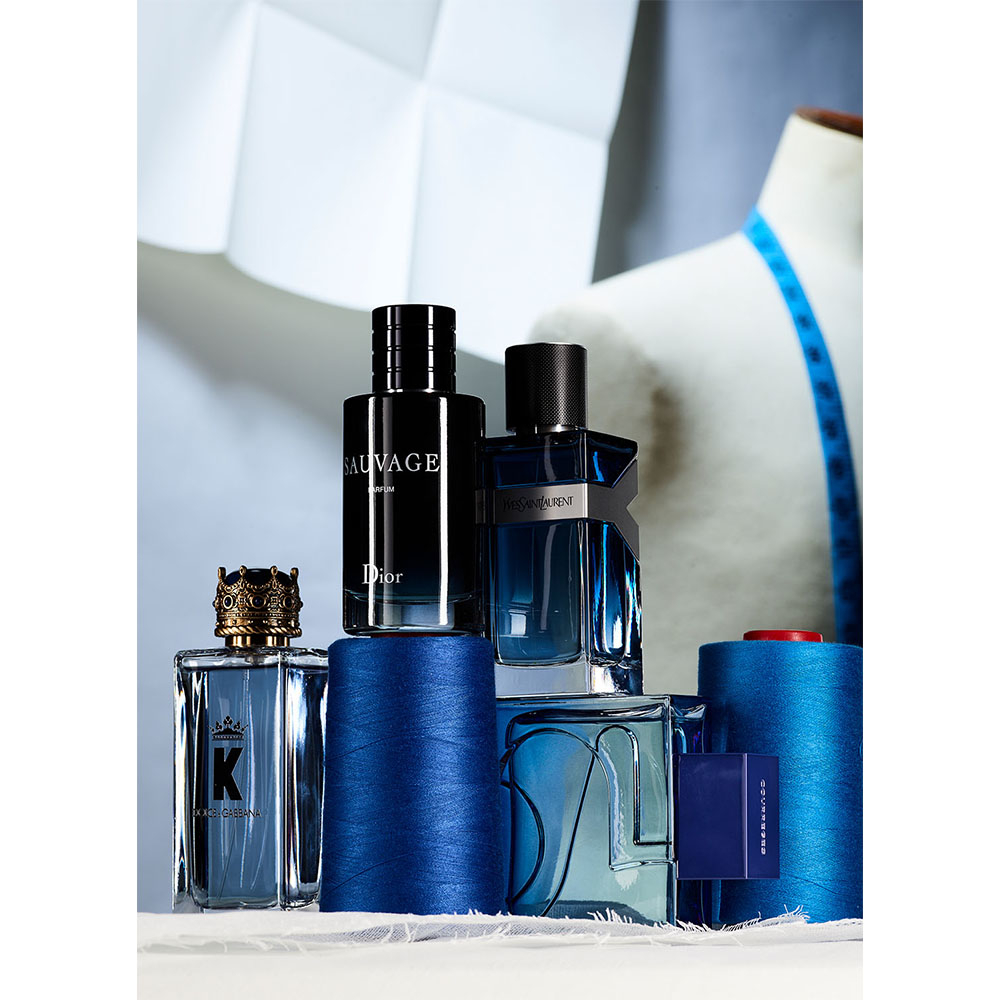A vibrant studio shot showcases an assortment of luxury perfume bottles arranged on spools of blue thread with tailoring mannequin in the backdrop.
