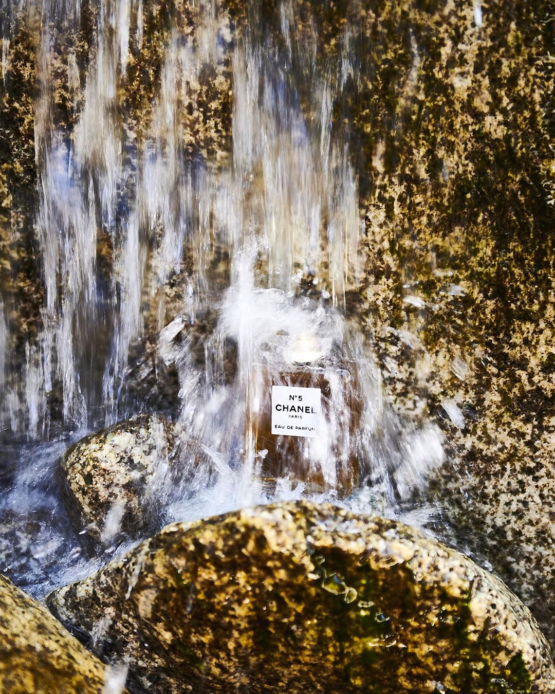 Chanel N°5 perfume bottle on stones with water cascading, beauty and fragrance, luxury and brand association, close-up shot.