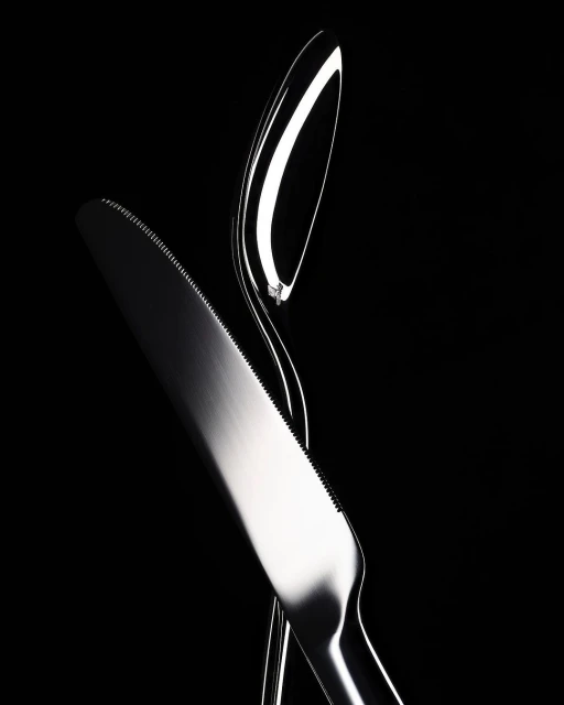 High contrast studio shot of a modern, shiny silver metal knife and spoon intersecting against a stark black background.