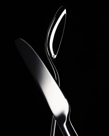 High contrast studio shot of a modern, shiny silver metal knife and spoon intersecting against a stark black background.
