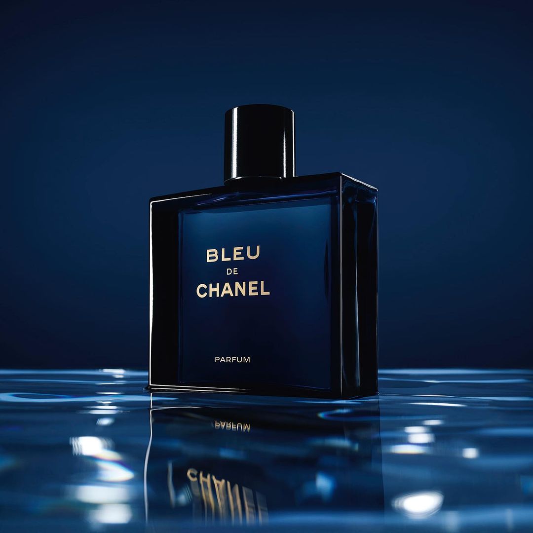 Square blue glass parfum bottle of Bleu de Chanel with black cap sits on reflective water-like surface against a blue background.
