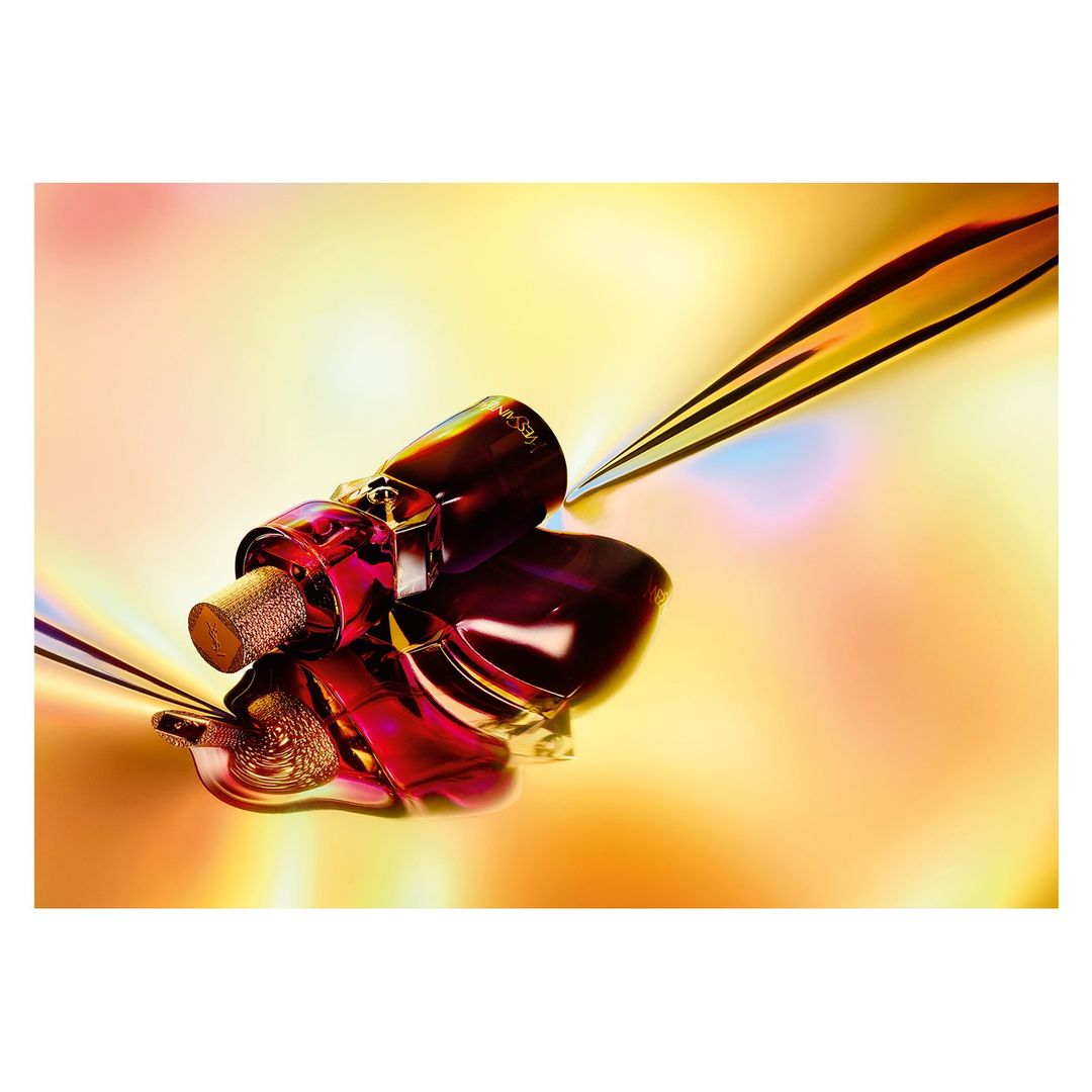 Still life close-up of a glossy, angular perfume bottle with a textured cork stopper on a warm-toned, iridescent background.