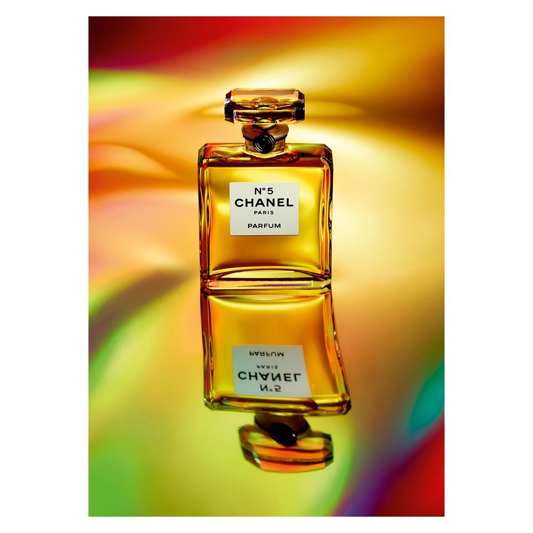Chanel N°5 perfume bottle on glossy surface reflects vibrant gradient background, elegant studio shot for beauty and fashion.