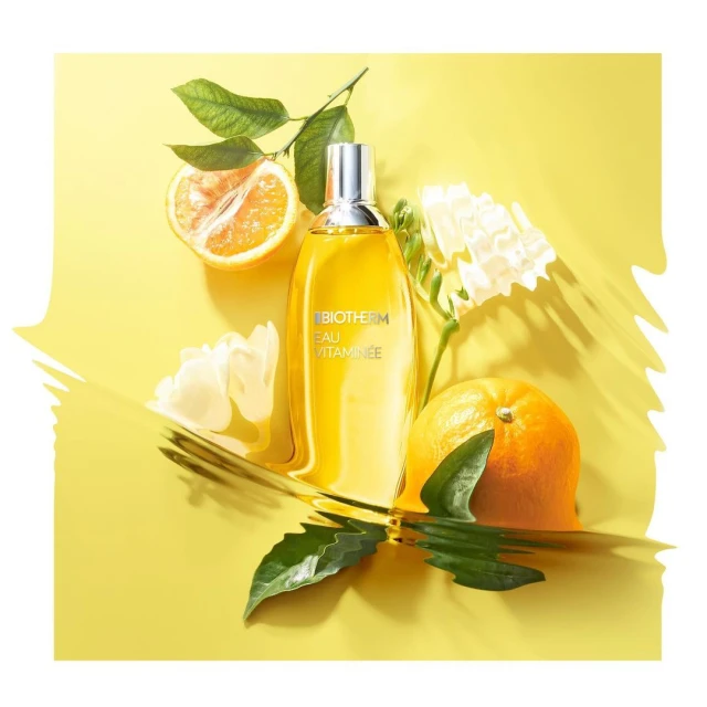 Bottle of Biotherm Eau Vitaminée surrounded by citrus fruits and white flowers on a yellow background, product photography.