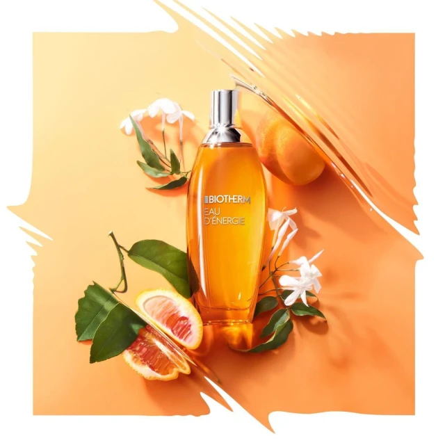 Beauty shot of Biotherm Eau d'Énergie fragrance bottle surrounded by grapefruit and jasmine flowers on a light orange backdrop.