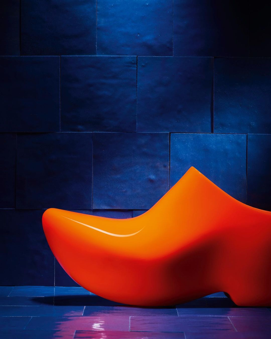Abstract studio shot of a large, orange, shoe-shaped chair against a blue tiled wall with a matching blue reflective floor.