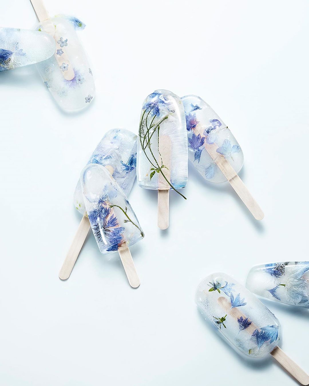Several ice pops with edible flowers frozen inside are lying on a white surface under bright lighting with a shallow depth of field.