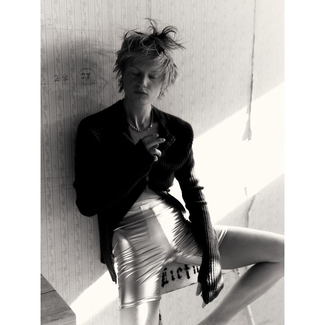 Black and white portrait of a young woman in a cardigan and shiny shorts leaning against a wall with peeling wallpaper indoors.