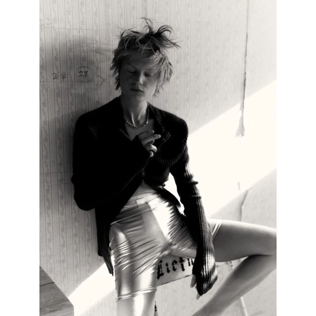 Black and white portrait of a young woman in a cardigan and shiny shorts leaning against a wall with peeling wallpaper indoors.