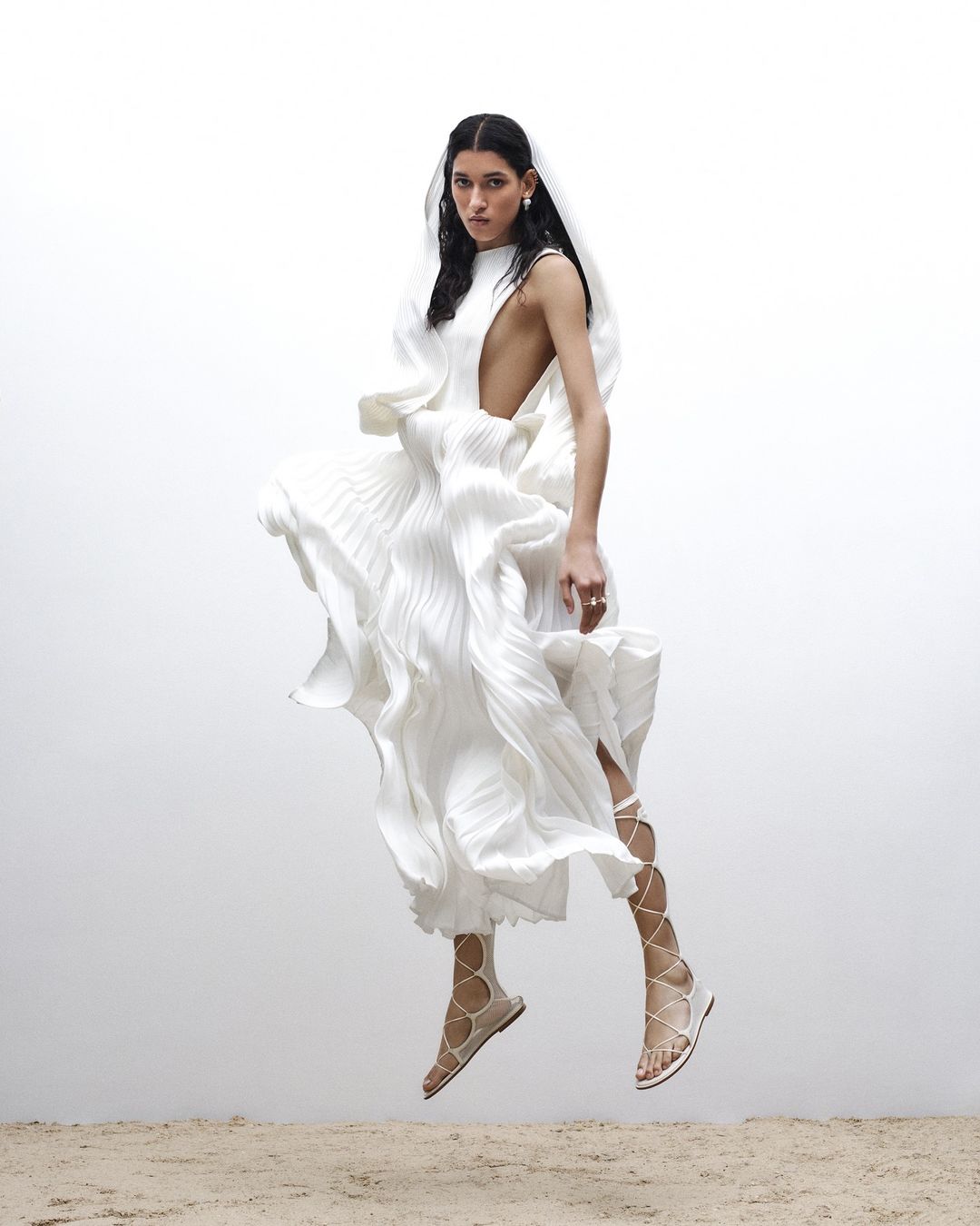 Fashion shot of a female model in a flowing white dress and gladiator sandals, elevated above a sandy surface against a bright background.
