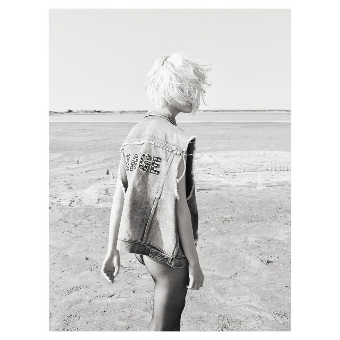 Back view of a person with blonde hair in a denim vest standing on a sandy beach with a body of water in the distance.