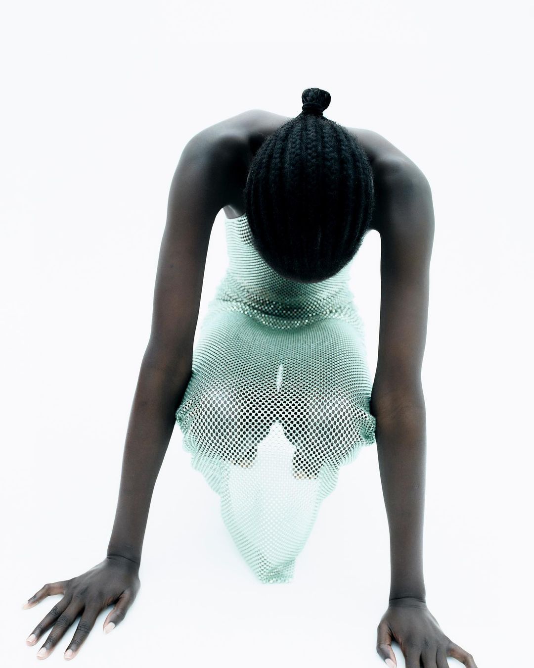 A person with dark skin and a bun is leaning forward with straight arms wearing a see-through dress against a bright white background.