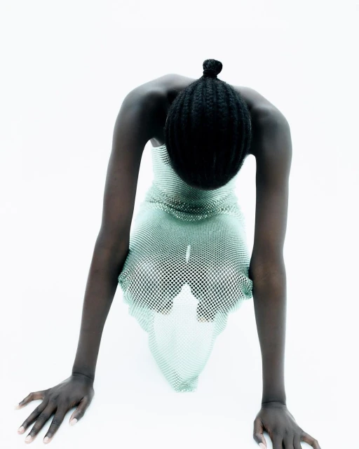 A person with dark skin and a bun is leaning forward with straight arms wearing a see-through dress against a bright white background.
