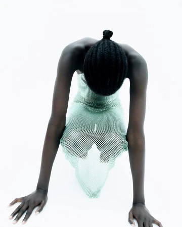 A person with dark skin and a bun is leaning forward with straight arms wearing a see-through dress against a bright white background.