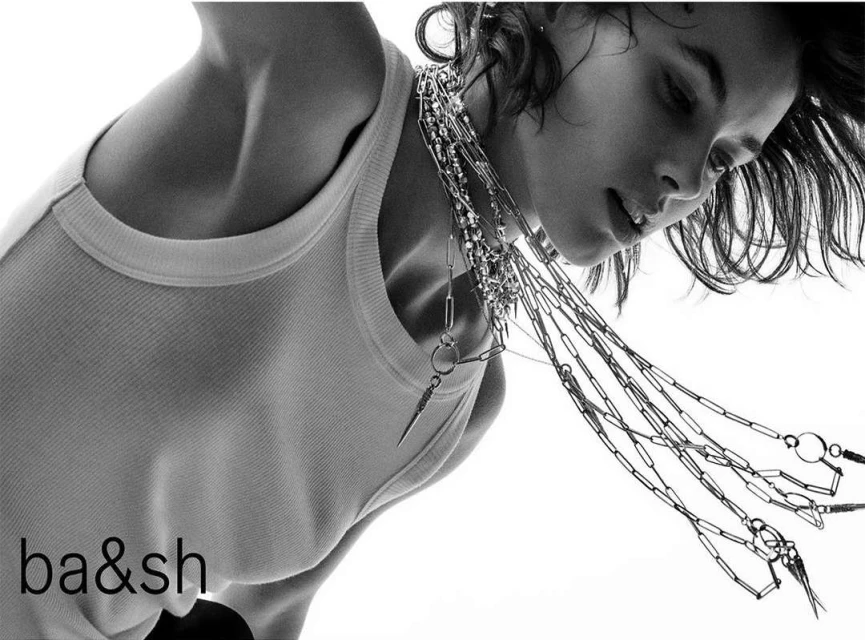 Monochrome studio shot of a young adult woman wearing a white tank top and layered chain necklaces looking down against a white backdrop.
