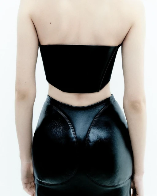 Back view of a slender adult female with light skin in a black strapless top and shiny black skirt against a white backdrop.