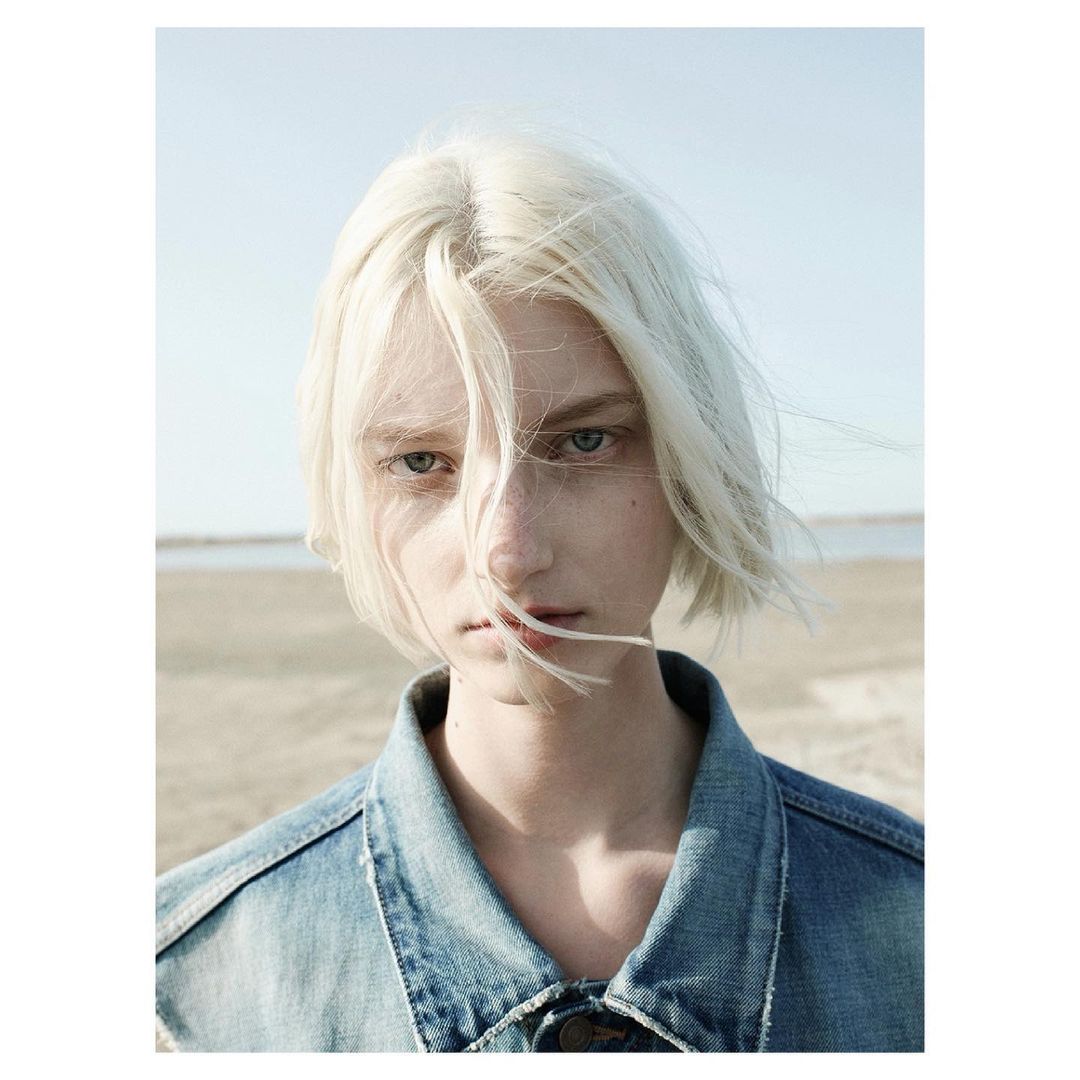 Portrait of a person with short, platinum blonde hair and blue eyes, wearing a blue denim jacket, standing outdoors with a neutral expression.