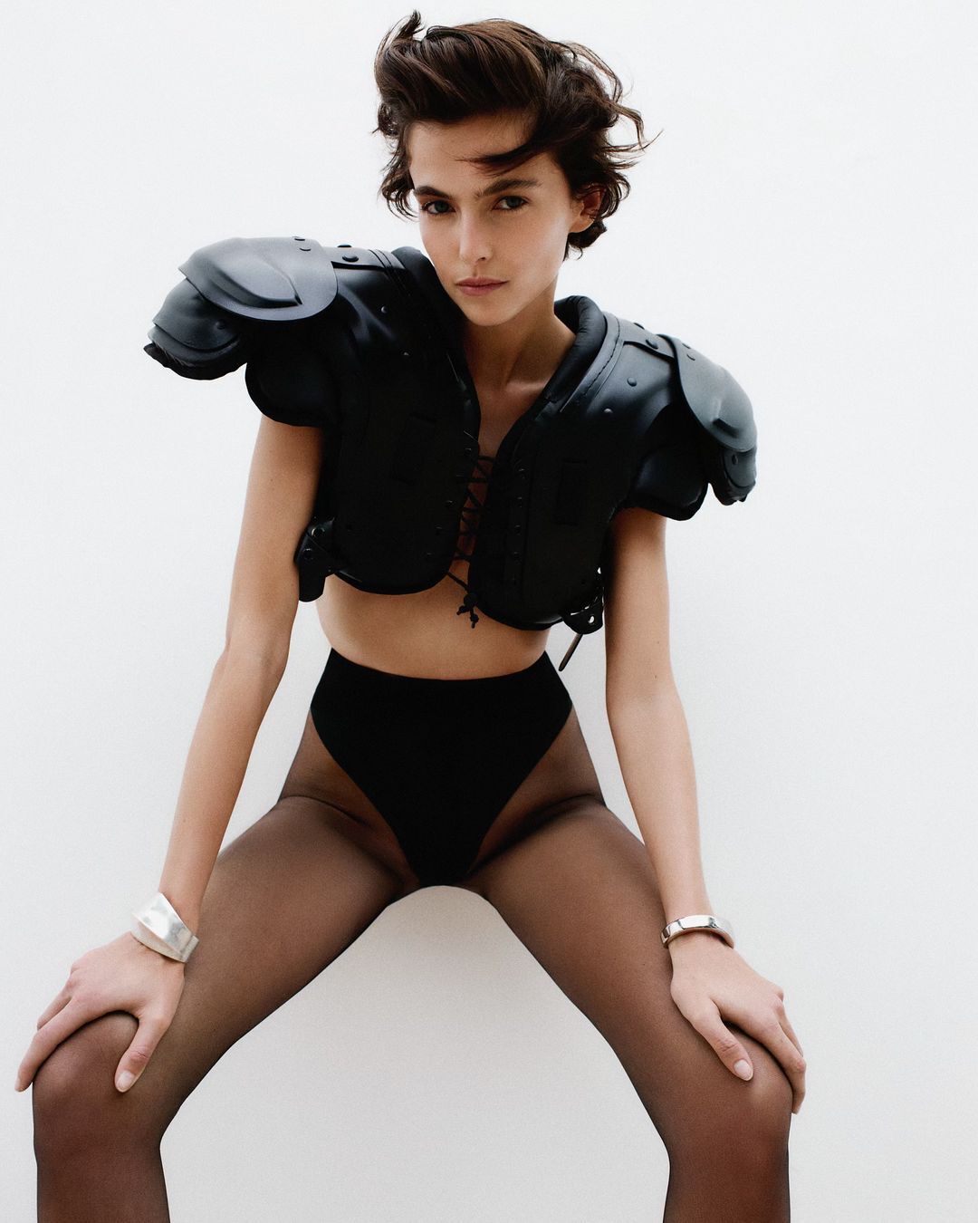 Fashionable adult woman poses in high-waisted black bottoms, sheer black tights, and football shoulder pads against a white backdrop.