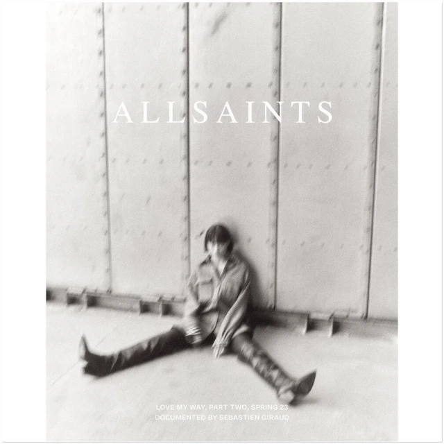 Monochrome image featuring a single adult sitting in front of a textured wall, promoting AllSaints Spring 23 collection with diffused lighting.