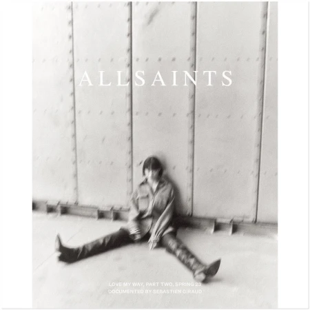 Monochrome image featuring a single adult sitting in front of a textured wall, promoting AllSaints Spring 23 collection with diffused lighting.