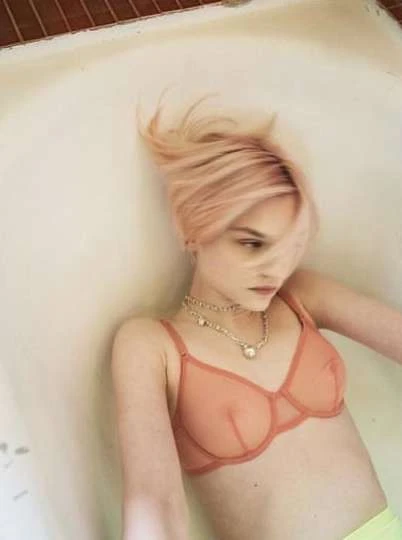 A woman with pink hair is lying in a bathtub, wearing a bra and necklace. Eye-level shot of a person against a neutral-toned background.